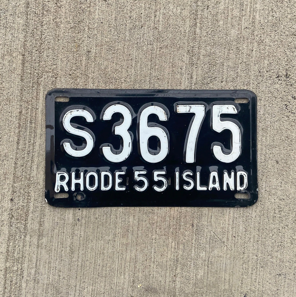 Photo of a 1955 Rhode Island license plate on a concrete background.