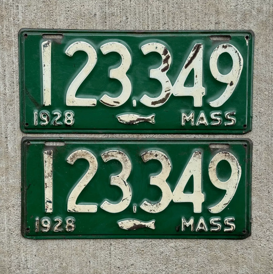 Pair of vintage 1928 Massachusetts license plates number 123359 with iconic cod fish graphic. Plates displayed on a concrete background.