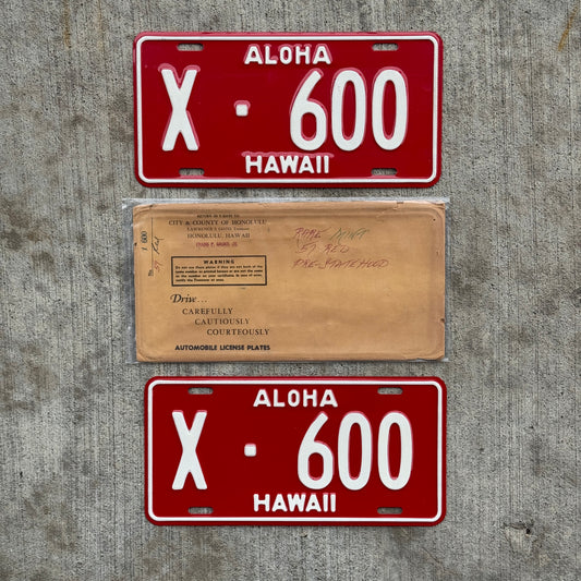Photo of a vintage 1957 Hawaii License Plate Pair Auto Tag New Old Stock Honolulu X 600 on a concrete background with their original mailing envelope.