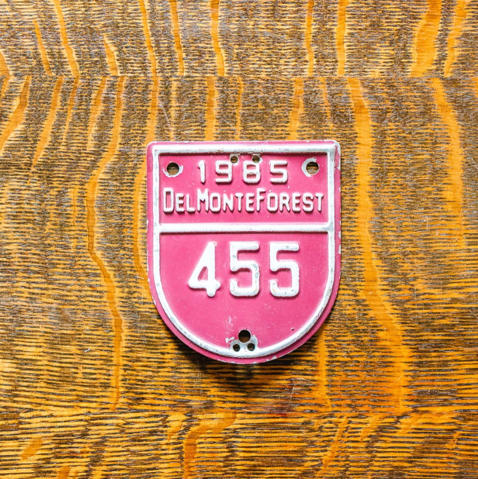 Red and white vintage-style license plate with '1985 Del Monte Forest 455' text on a wooden surface.